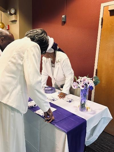 Guest in white attire signing in at the registration table with purple runner and calla lily centerpiece — Golden Jubilee All-White Affair 2026