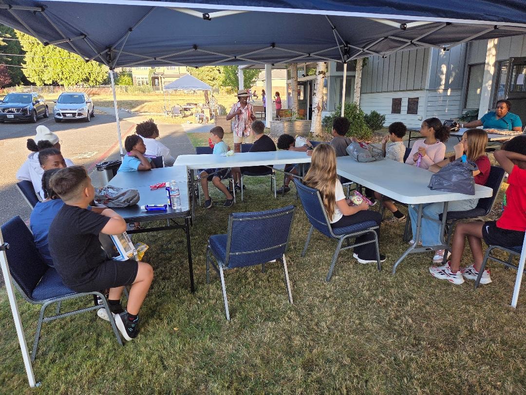 Children seated at outdoor tables under tent — VBS Day 3 Assembly