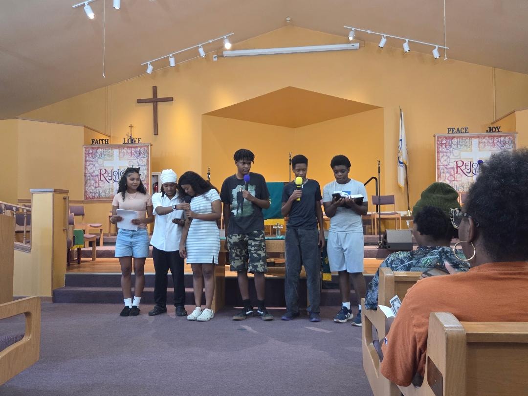 Two women seated in pews with speaker at podium — VBS Day 3 Assembly, MLK Memorial Baptist Church