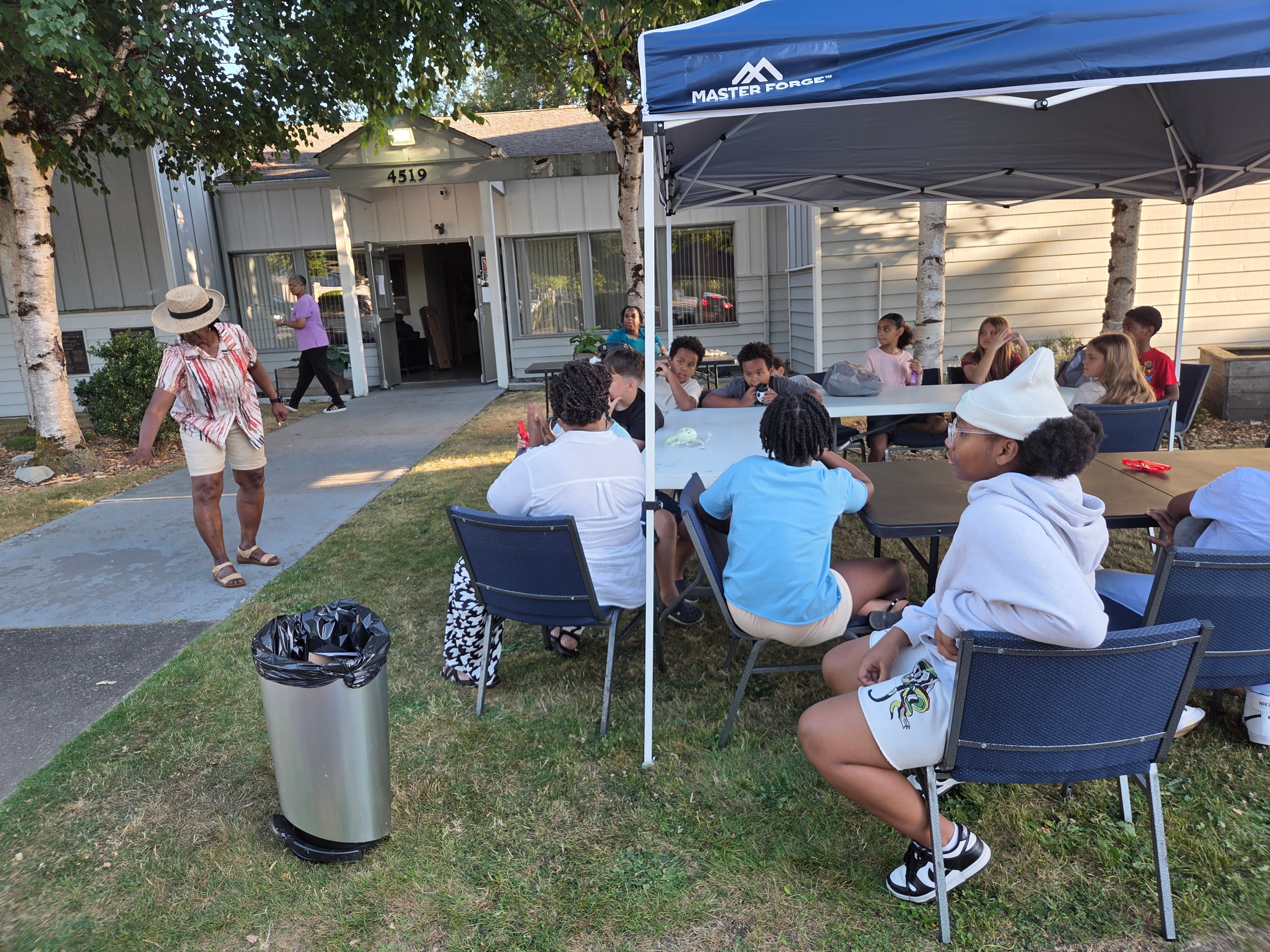 Children seated at tables under blue tent outside church — VBS Day 3 Assembly