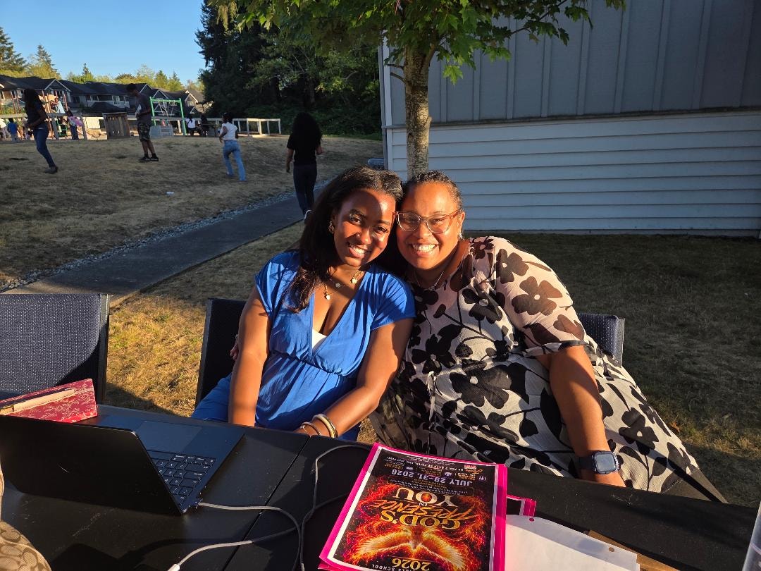 Two women smiling together at outdoor table with VBS program — VBS Day 3 Assembly