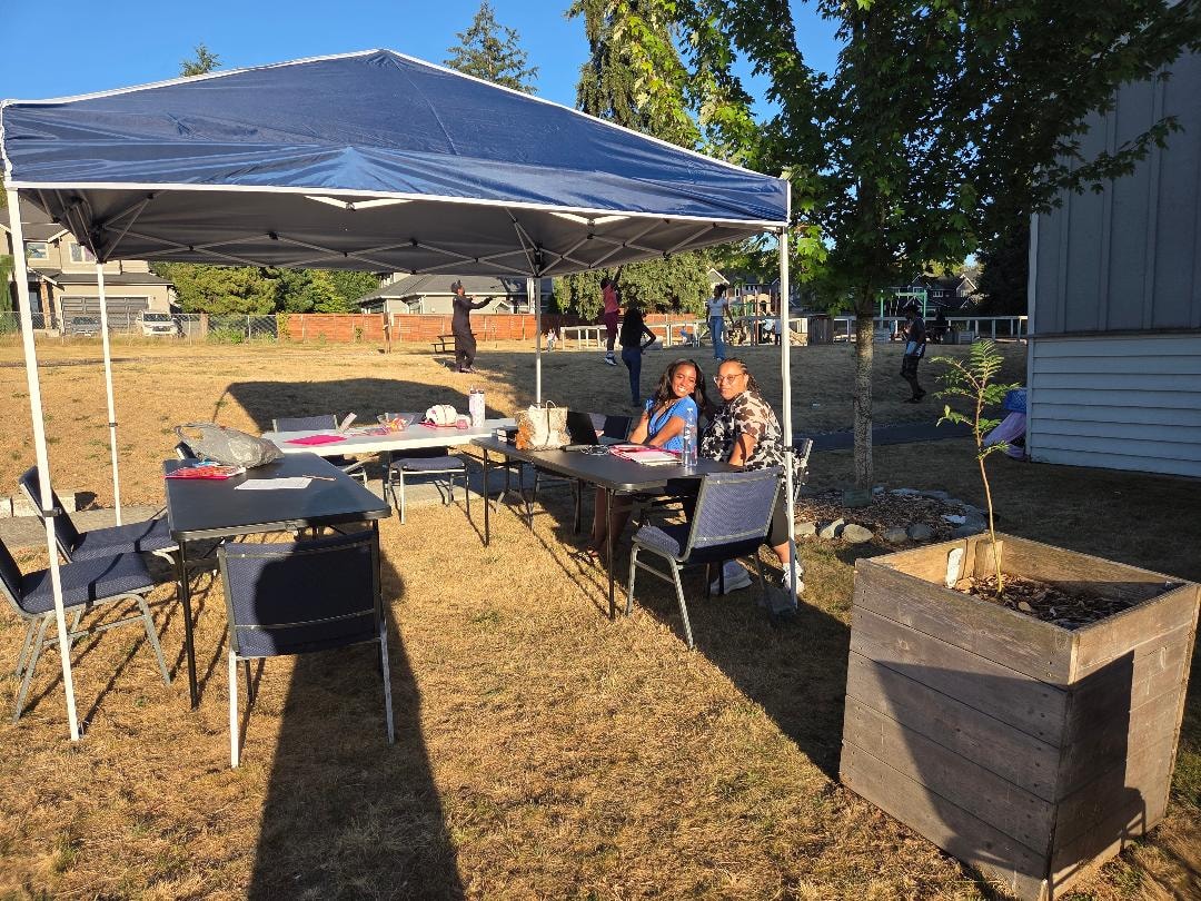 Two girls seated at table under blue tent on church grounds — VBS Day 3 Assembly